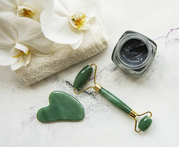 Πρωινή ρουτίνα ομορφιάς skincare στο σπίτι μπάνιο - πρόσωπο νεφρίτη roller massager για wellness καλλυντική θεραπεία, μάσκα και ορχιδέα λουλούδια σε μαρμάρινο φόντο - Φωτογραφία, εικόνα