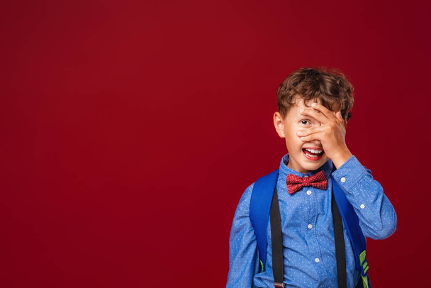 Portrait of a schoolboy, a Caucasian boy with wavy hair in a uniform and with a backpack. made a mistake, covers his face with his hand and looks through his fingers. Posing on a red background. - Fotó, kép