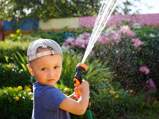 ein kleines Kind in einer Mütze, das die Pflanzen mit einem Schlauch wässert 2024 - Foto, Bild