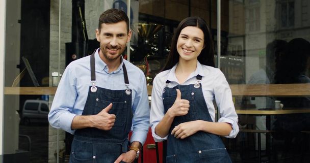 Young Caucasian cheerful waiter and waitress turning to the camera, giving their thumbs up and smiling while standing at the entrance to the restaurant. Portrait shot. - Photo, image