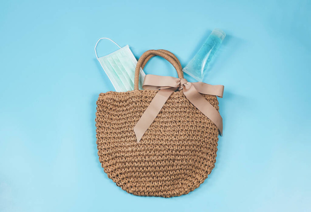 COVID-19 prevention, travel and new normal concept, top view of woven bag with sanitizer gel and surgical face mask on blue background. - Фото, изображение