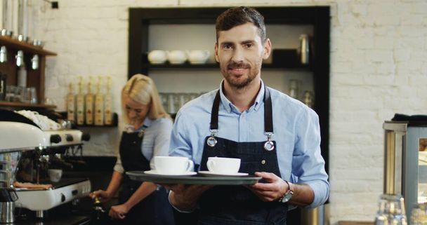 Portrait du beau barmen caucasien tenant des tasses de café et allant au bar tandis que sa collègue féminine se prépare plus derrière à la machine à café. - Photo, image