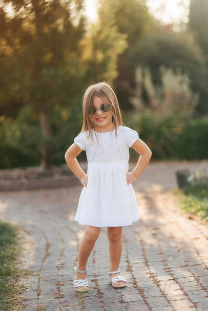 Roztomilá holčička v bílých šatech pózující fotografce. Šťastné dítě ve slunečních brýlích - Fotografie, Obrázek