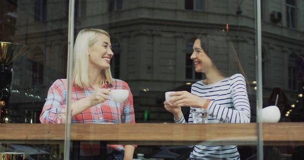 Two smiled pretty female friends sitting in the cafe behind window, taking nicely and drinking coffee. - Foto, Imagen