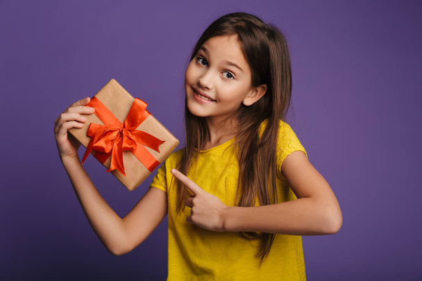Photo of happy beautiful girl holding and pointing finger at gift box isolated over purple background - Φωτογραφία, εικόνα