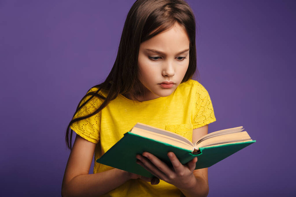 Photo of smart focused girl posing and reading book on camera isolated over purple background - Photo, Image