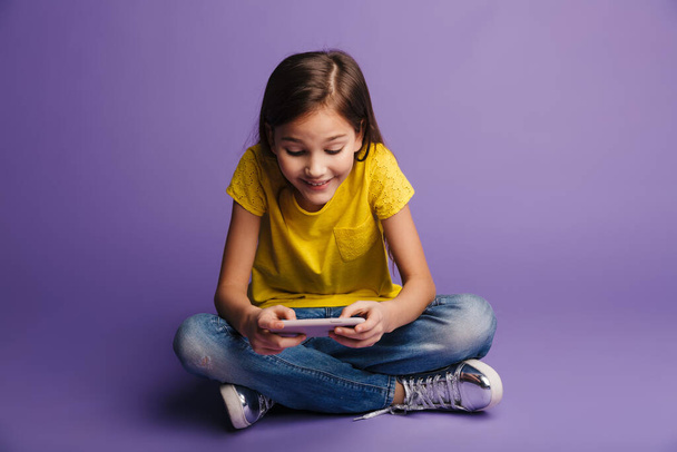 Photo of smiling cute girl playing video game on cellphone while sitting on floor isolated over purple background - 写真・画像
