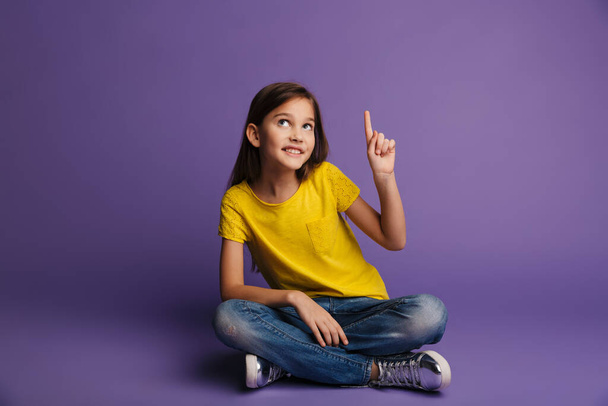 Photo of smiling cute girl pointing finger upward while sitting on floor isolated over purple background - Zdjęcie, obraz