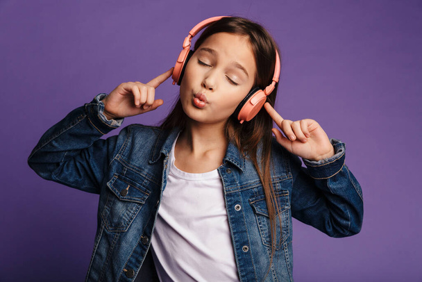 Photo de gentille fille gesticulant la paix chantant et utilisant des écouteurs sans fil isolés sur fond violet - Photo, image