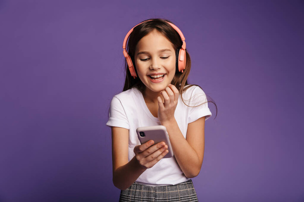Photo of laughing attractive girl using wireless headphones and mobile phone isolated over purple background - Zdjęcie, obraz