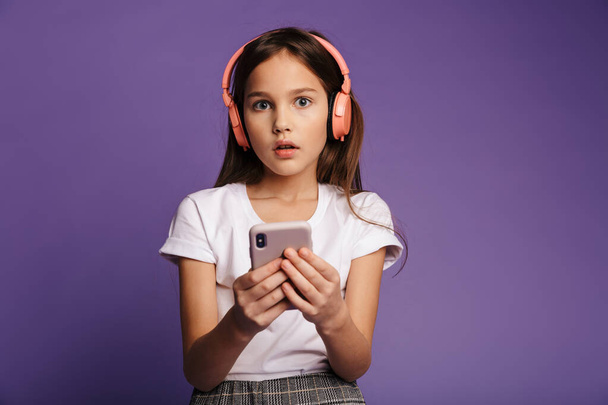 Photo of confused attractive girl using wireless headphones and mobile phone isolated over purple background - Foto, afbeelding
