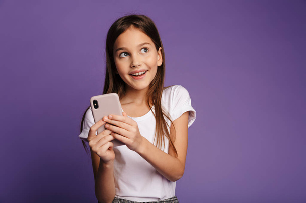Fotografie usměvavé atraktivní dívka pomocí mobilního telefonu a při pohledu stranou izolované přes fialové pozadí - Fotografie, Obrázek