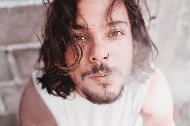 Young dark-haired attractive sexy mixed-race man smokes a cigarette, blows smoke. close-up portrait. Beauty and fashion people. The harm and aesthetics smoking. - 写真・画像