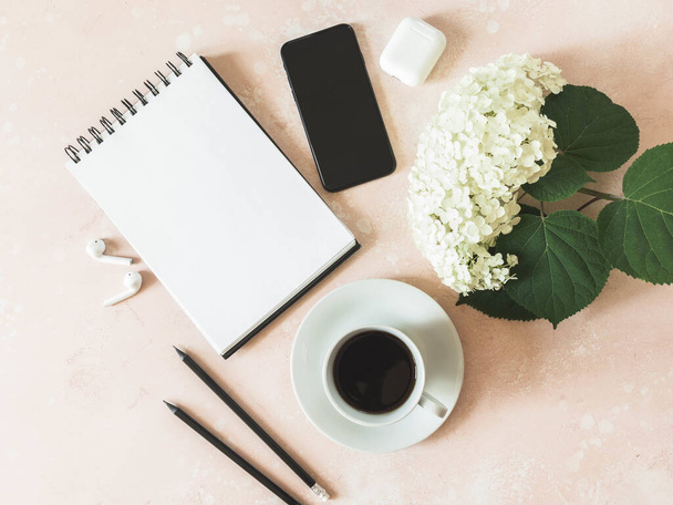 Home office desk workspace with black smartphone, notepad, black coffee in cup, white hydrangea, white earphones and graphite pencils on pink background. Flat lay, top view girl work business concept for lifestyle blog - Фото, изображение