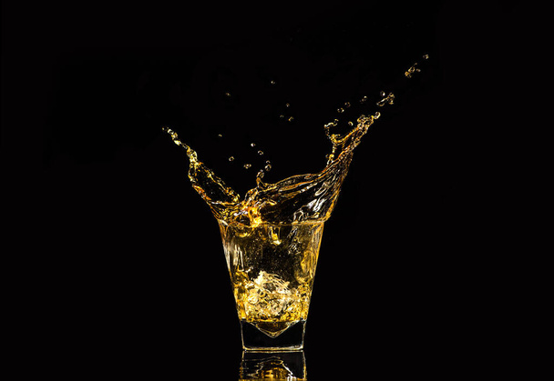 Ένα ποτήρι ουίσκι με πιτσιλιές από το παγάκι πάνω σε μαύρο φόντο. Πιτσιλιές αλκοόλ. ουίσκι ή κονιάκ ή άλλο είδος αλκοόλ με πιτσιλιές. - Φωτογραφία, εικόνα