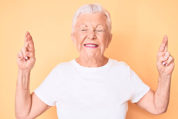 Senior mooie vrouw met blauwe ogen en grijs haar dragen klassieke witte tshirt over gele achtergrond gebaren vinger gekruist glimlachen met hoop en ogen dicht. geluk en bijgelovig concept.  - Foto, afbeelding
