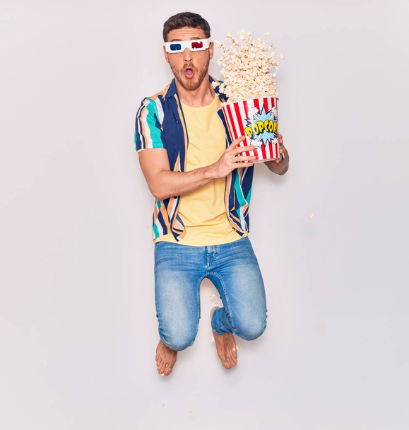 Jonge knappe Spaanse man verrast met een open mond met een 3d bril. emmer popcorn die over een geïsoleerde witte achtergrond springt. - Foto, afbeelding