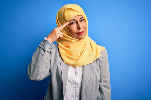 Donna d'affari bruna di mezza età che indossa hijab tradizionale musulmano su sfondo blu che indica infelice brufolo sulla fronte, brutta infezione di blackhead. Acne e problemi della pelle - Foto, immagini