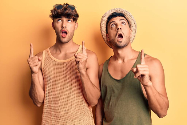 Молодая пара геев в летней одежде удивлена и удивлена, глядя вверх и указывая пальцами и подняв руки.  - Фото, изображение