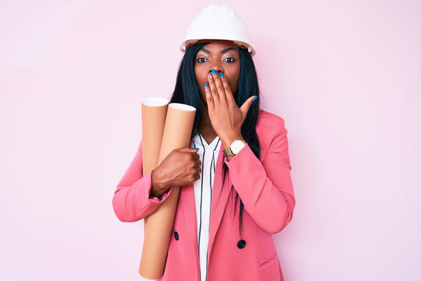 Jonge Afro-Amerikaanse vrouw met een architectenhoed met blauwdrukken die mond met hand bedekken, geschokt en bang voor fouten. verraste uitdrukking  - Foto, afbeelding