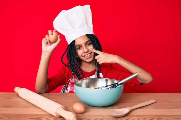 Junge afrikanisch-amerikanische Mädchen mit Zöpfen tragen professionelle Kochschürze hält Ei lächelnd glücklich zeigt mit Hand und Finger  - Foto, Bild