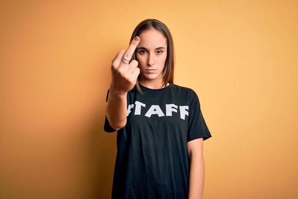 Young beautiful worker woman wearing staff uniform t-shirt over isolated yellow background Showing middle finger, impolite and rude fuck off expression - Фото, изображение