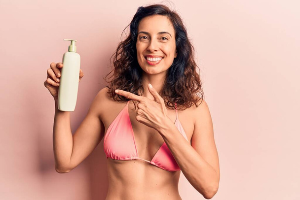Jonge mooie Spaanse vrouw draagt bikini met zonnebrandcrème en glimlacht vrolijk wijzend met hand en vinger  - Foto, afbeelding