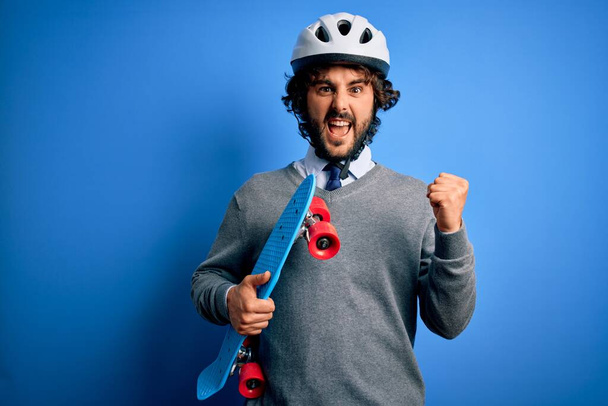髭を生やしたハンサムなスケーター男は青い背景の上にスケートを保持しているセキュリティヘルメットを身に着けている誇りと勝利と成功を祝って叫んで非常に興奮し、応援感情 - 写真・画像