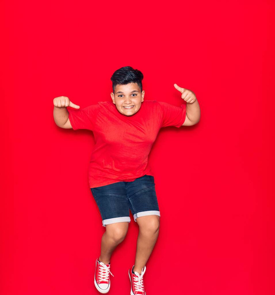 Adorable kid wearing casual clothes smiling happy. Jumping with smile on face doing ok sign with thumbs up over isolated red background - Photo, image