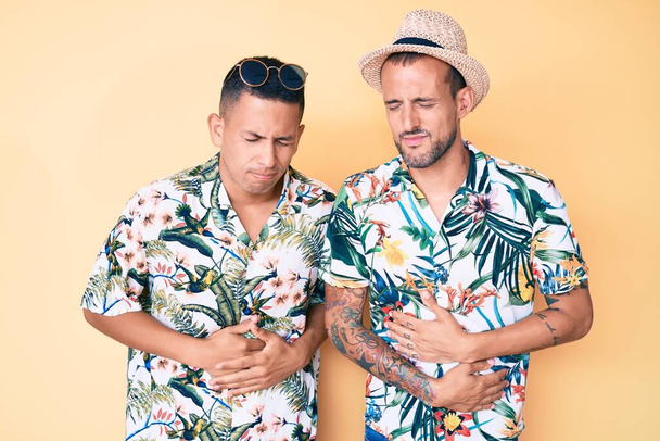 Jeune couple gay de deux hommes portant un chapeau d'été et chemise hawaïenne avec la main sur le ventre parce que l'indigestion, la maladie douloureuse se sent mal. concept de douleur.  - Photo, image