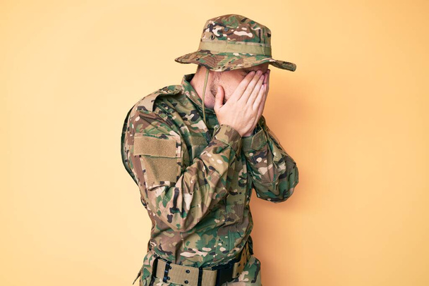 Giovane uomo caucasico con uniforme militare mimetica con espressione triste che copre il viso con le mani mentre piange. concetto di depressione.  - Foto, immagini