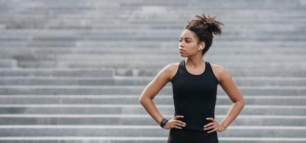Ćwiczenia joggingowe i zdrowy tryb życia. Poważnie afrykańska amerykańska dziewczyna w sportowej odzieży z nadajnikiem fitness na szarym tle schodów - Zdjęcie, obraz