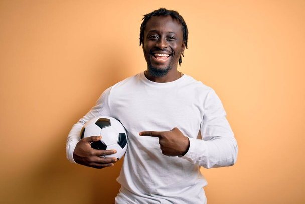 若いですアフリカ系アメリカ人選手男遊びサッカーボールを持っています黄色の背景上に非常に幸せなポインティングとともに手と指 - 写真・画像