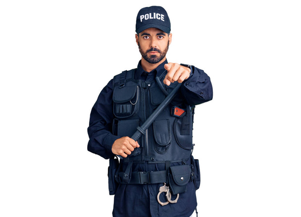 Jeune homme hispanique portant l'uniforme de police tenant bâton pointant du doigt la caméra et à vous, geste confiant regardant sérieux  - Photo, image