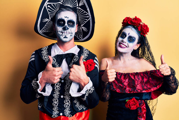 Ölü Meksika kostümlü genç bir çift sarı başarı işaretinin üzerinde el hareketleriyle pozitif jest yapıyor, gülümsüyor ve mutlu. Neşeli ifade ve kazanan jest.  - Fotoğraf, Görsel