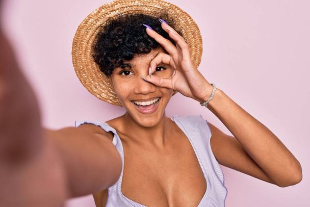 Όμορφη Αφροαμερικάνα τουρίστρια γυναίκα φορώντας μπικίνι και καπέλο κάνοντας selfie με κάμερα με χαρούμενο πρόσωπο χαμογελώντας κάνει ok σημάδι με το χέρι στο μάτι κοιτάζοντας μέσα από τα δάχτυλα - Φωτογραφία, εικόνα