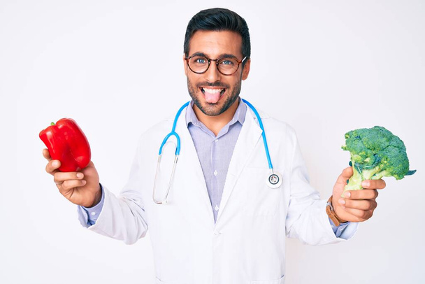 Young hispanic man wearing doctor stethoscope holding red pepper and broccoli sticking tongue out happy with funny expression.  - Photo, Image