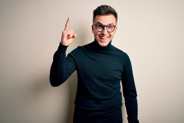 Junger gutaussehender kaukasischer Mann mit Brille und lässigem Pullover vor isoliertem Hintergrund, der mit erhobenem Zeigefinger auf eine erfolgreiche Idee hinweist. Aufgeregt und glücklich. Nummer eins. - Foto, Bild