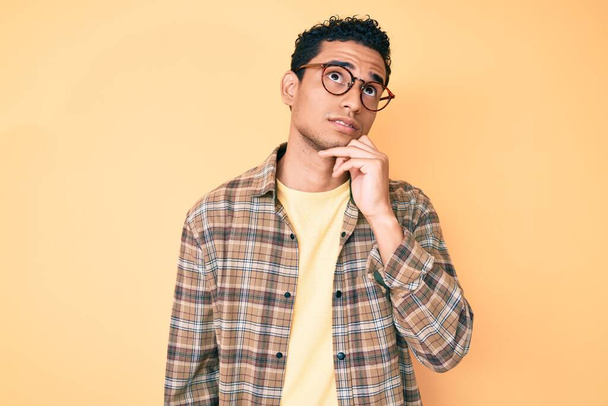 Young handsome hispanic man wearing casual clothes and glasses serious face thinking about question with hand on chin, thoughtful about confusing idea  - Foto, immagini