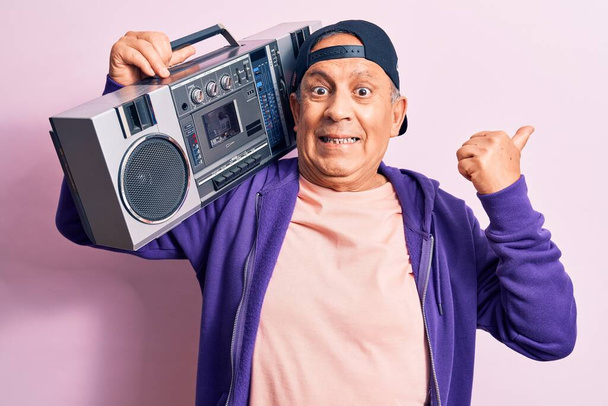 Senior handsome grey-haired modern man listening to music using vintage boombox pointing thumb up to the side smiling happy with open mouth - Фото, изображение