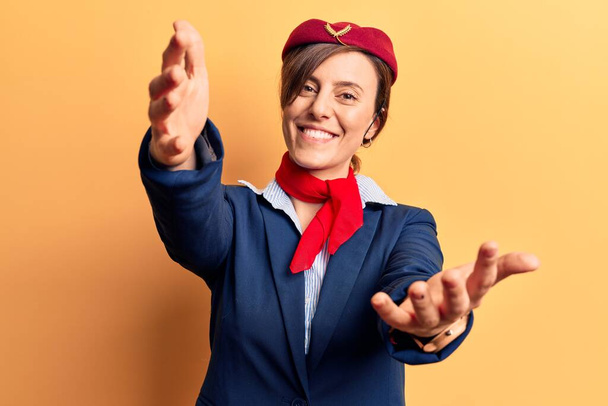 Junge schöne Frau in Stewardess-Uniform, die in die Kamera lächelt und mit offenen Armen umarmt wird. fröhlicher Ausdruck, der das Glück umarmt.  - Foto, Bild