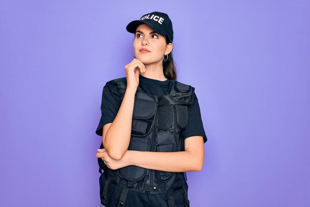Egy fiatal rendőrnő golyóálló mellényt visel lila háttér felett, kezével az állán, gondolkodva a kérdésen, töprengő arckifejezéssel. Mosolygó és figyelmes arc. Kétséges fogalom. - Fotó, kép