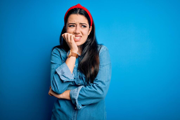 Jonge brunette vrouw draagt casual denim shirt over blauwe geïsoleerde achtergrond kijken gestrest en nerveus met handen op de mond bijtende nagels. Angst probleem. - Foto, afbeelding