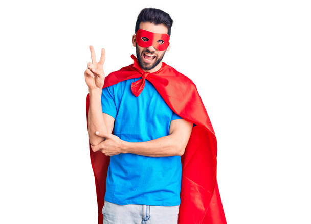Young handsome man with beard wearing super hero costume smiling with happy face winking at the camera doing victory sign. number two.  - Foto, immagini
