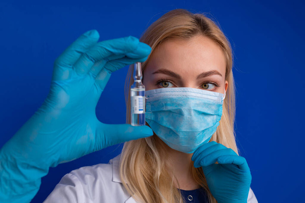 Tıbbi maskeli ve eldivenli bir kız doktor mavi arka planda hastalığa karşı aşısı olan bir ampul tutuyor. - Fotoğraf, Görsel