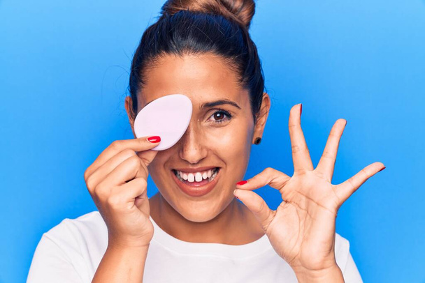 Young beautiful brunette woman holding makeup sponge doing ok sign with fingers, smiling friendly gesturing excellent symbol  - Photo, Image