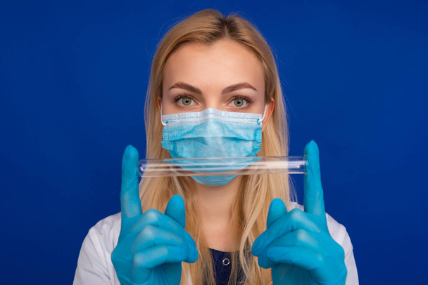 A girl doctor in a medical mask and gloves holding test tube on a blue background - Foto, afbeelding