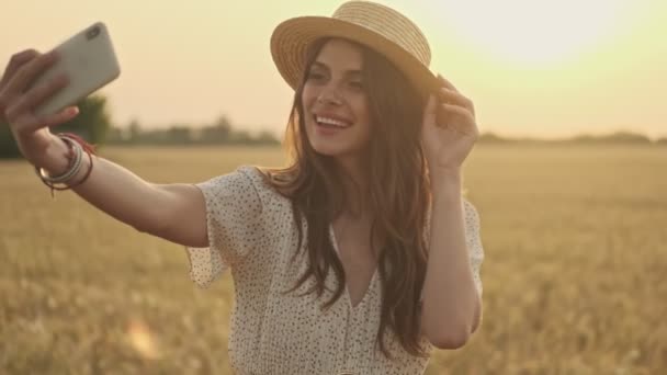 Happy pretty woman wearing dress and stylish hat making selfie on smartphone while posing on wheat field - Filmati, video