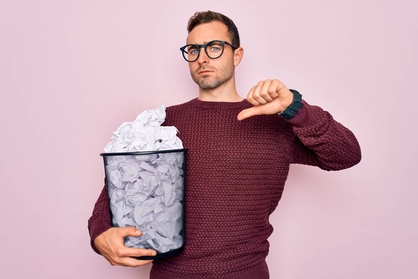 Handsome man with blue eyes wearing glasses holding full paper bin with crumpled papers with angry face, negative sign showing dislike with thumbs down, rejection concept - Foto, afbeelding