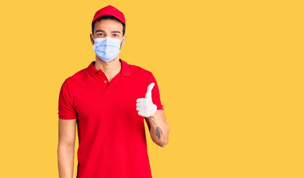 Young handsome hispanic man wearing delivery uniform and medical mask smiling happy and positive, thumb up doing excellent and approval sign  - Фото, зображення